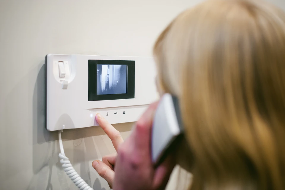 Femme blonde utilisant un interphone vidéo domestique, doigt pointé vers les commandes