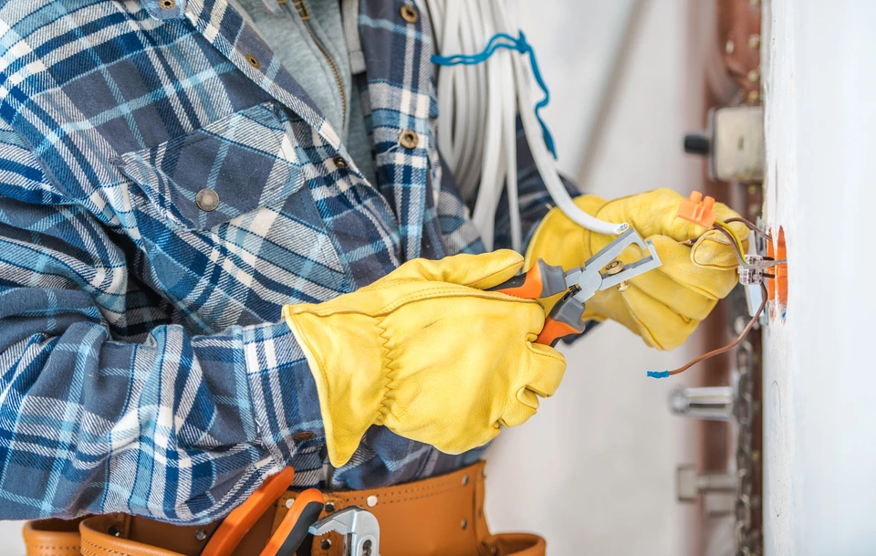 Électricien professionnel en chemise à carreaux travaillant sur installation électrique résidentielle, manipulant fils et connexions dans boîtier mural blanc