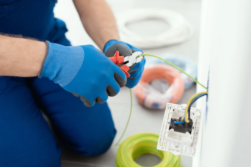 Électricien professionnel travaillant sur tableau électrique intérieur, portant lunettes de sécurité et casquette