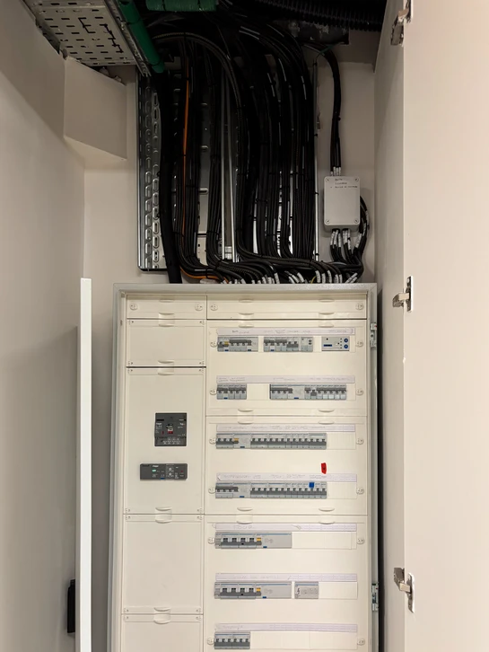 Photographie en couleur montrant un électricien professionnel en tenue de travail pratique (pantalon et chemise ou polo de travail), concentré et déterminé, intervenant sur un tableau électrique résidentiel. L'électricien tient un tournevis ou un testeur électrique, avec des outils visibles à proximité. L'arrière-plan montre l'intérieur d'une maison contemporaine. La scène doit transmettre professionnalisme, compétence technique et réactivité face à une situation d'intervention d'urgence. La lumière doit être naturelle et claire, typique d'un intérieur résidentiel.